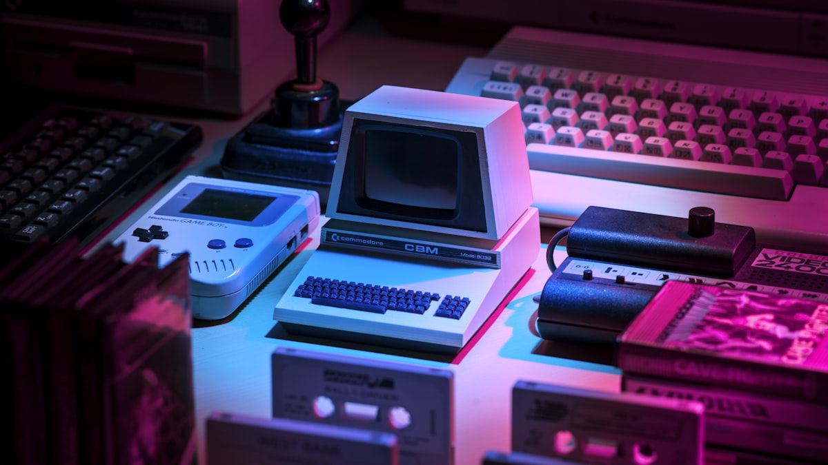 Vintage Commodore, Game Boy, and joystick in neon purple light — retro gaming hardware